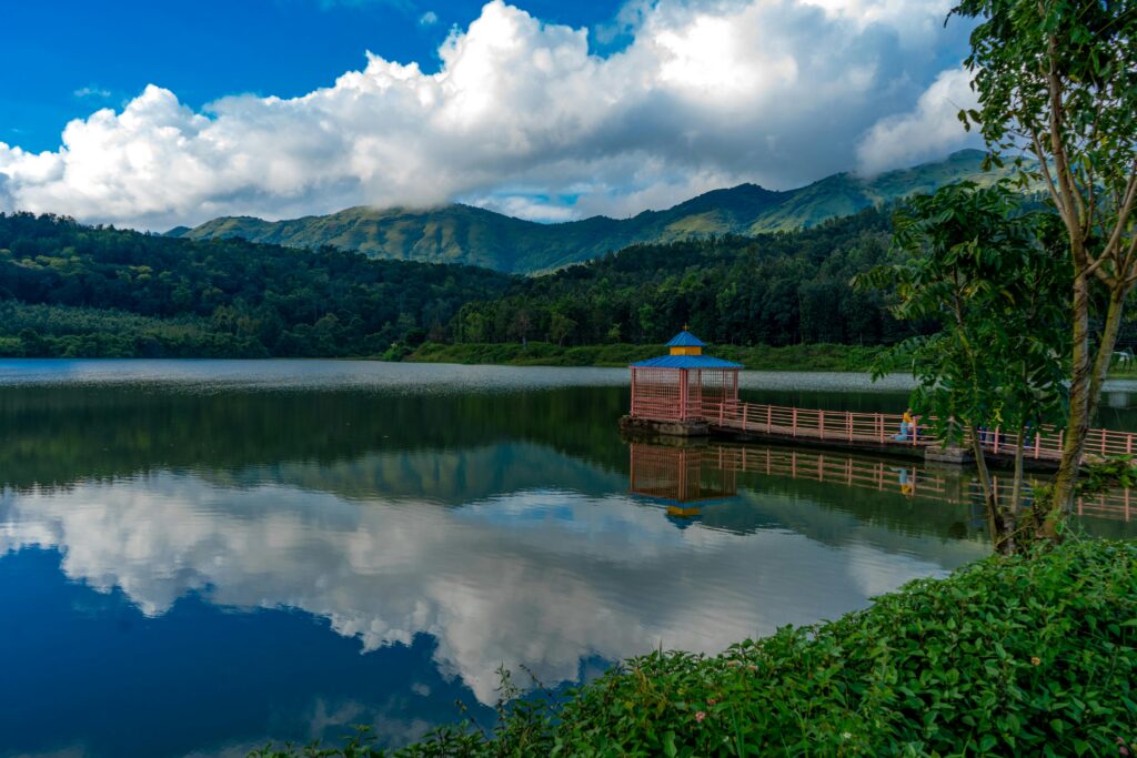 pexels-photo-10574299-10574299 Tranquil lake scene in HirekoLale, India, with a dock, lush greenery, and mountain backdrop under blue skies and fluffy clouds.