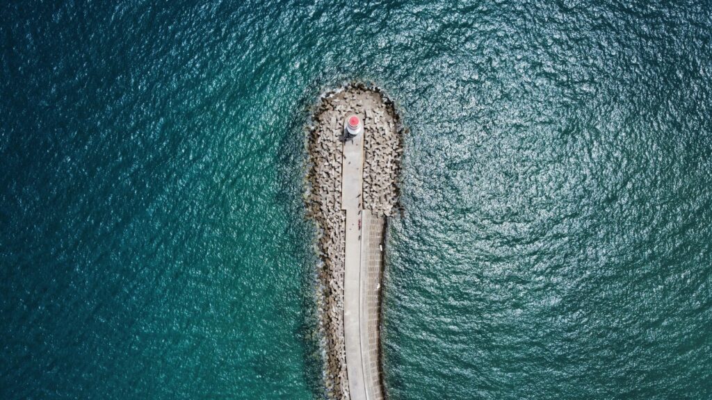 Aerial shot of a lighthouse on a jetty surrounded by clear blue ocean water, showcasing serene coastal beauty.