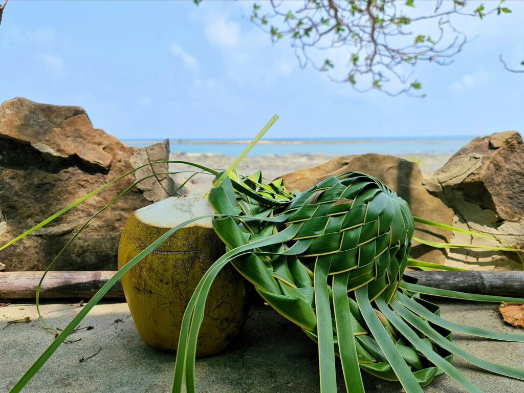 pexels-photo-30355247-30355247 A coconut, woven palm frond, rocks, and the ocean under a clear sky
