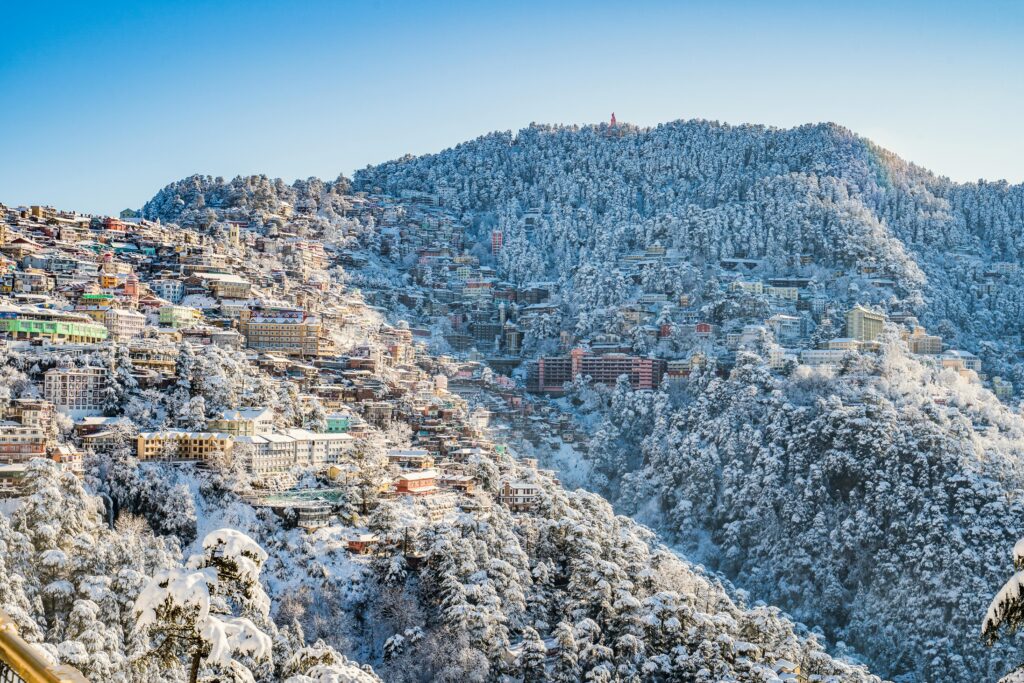 pexels-photo-3574440-3574440 A scenic view of Shimla in winter, with snow-covered buildings and forests under a clear blue sky.