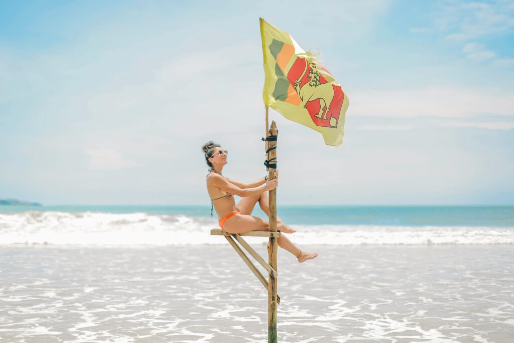pexels-photo-6337422-6337422 Woman in a bikini sitting on a wooden structure by the ocean with a Sri Lankan flag waving in the breeze.
